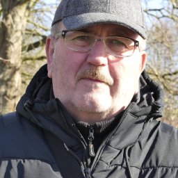 JOBB SERVICE D&W HB - Portret mężczyzny w czarnej kurtce i szarej czapce kaszkietówce na tle drzew w słoneczny dzień. Mężczyzna ma wąsy i okulary korekcyjne.