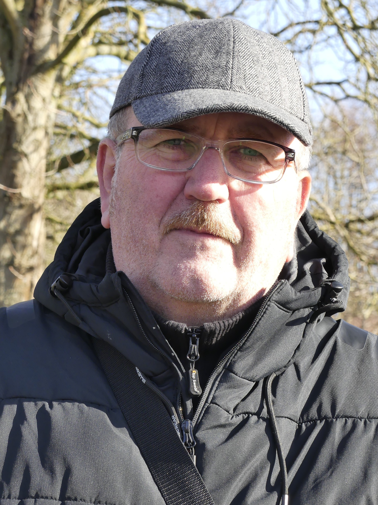 Portret mężczyzny w czarnej kurtce i szarej czapce kaszkietówce na tle drzew w słoneczny dzień. Mężczyzna ma wąsy i okulary korekcyjne.