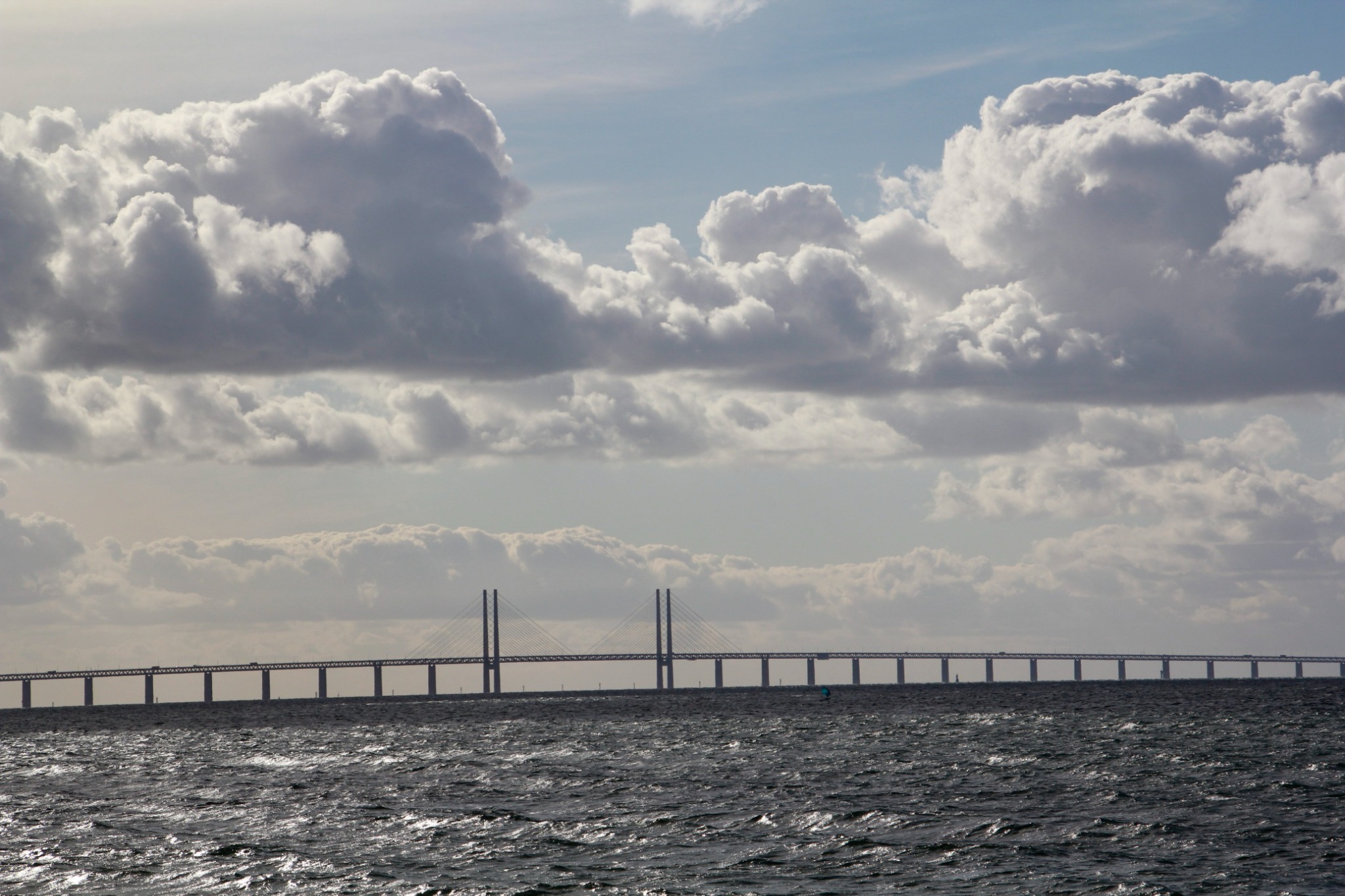 Długi most Øresund z dwoma pylonami, widziany z morza, pod pochmurnym niebem. Falująca woda odbija światło słoneczne.