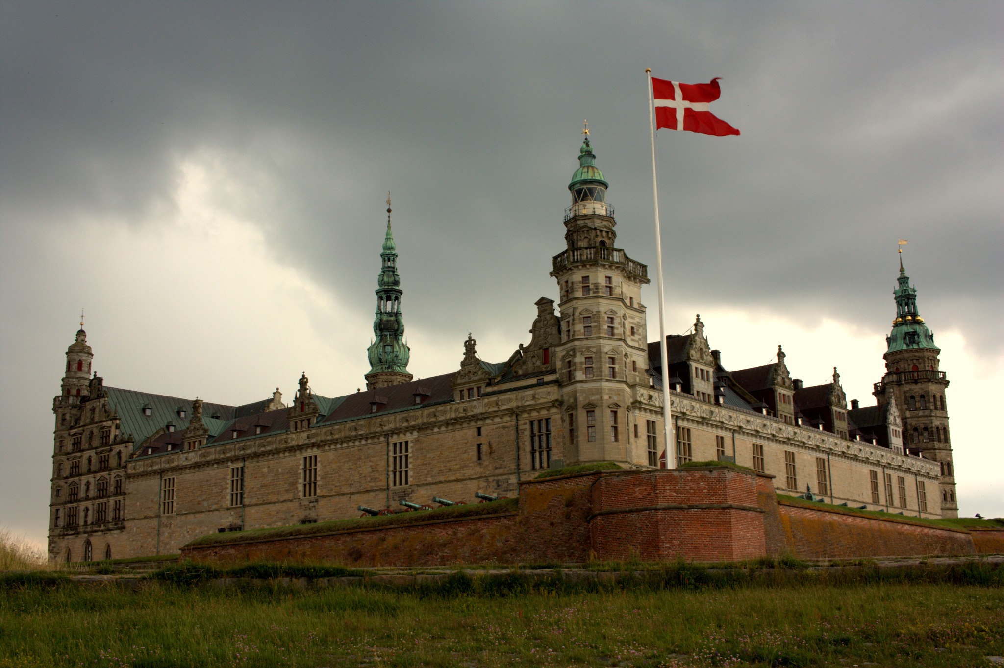 Królewski zamek Kronborg z powiewającą duńską flagą na tle pochmurnego nieba, widoczny z poziomu trawy. Na murach zamku ustawione są armaty.