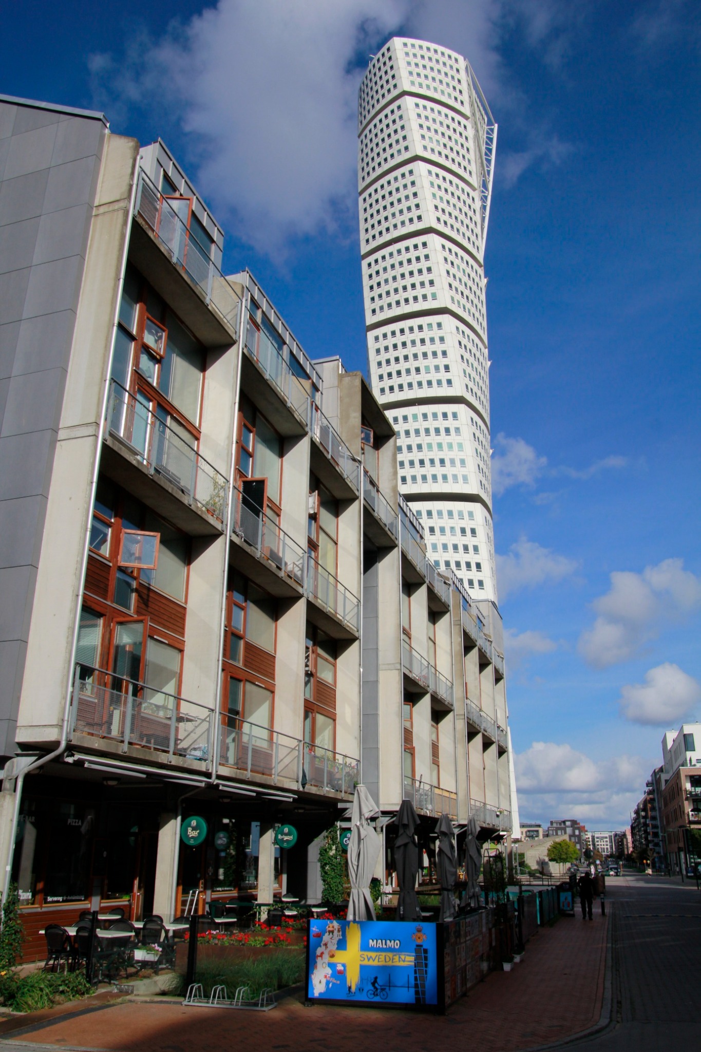 Architektura Malmö: Turning Torso góruje nad budynkiem z balkonami, w tle błękitne niebo z chmurami. Ulica z restauracją i dekoracyjnym panelem informacyjnym.
