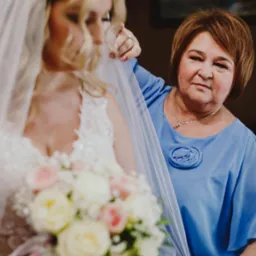 Panna młoda z bukietem, matka poprawia welon. Ujęcie z boku, ciepłe światło, widoczna faktura sukni i kwiatów. Kobieta w niebieskiej sukience z broszką.