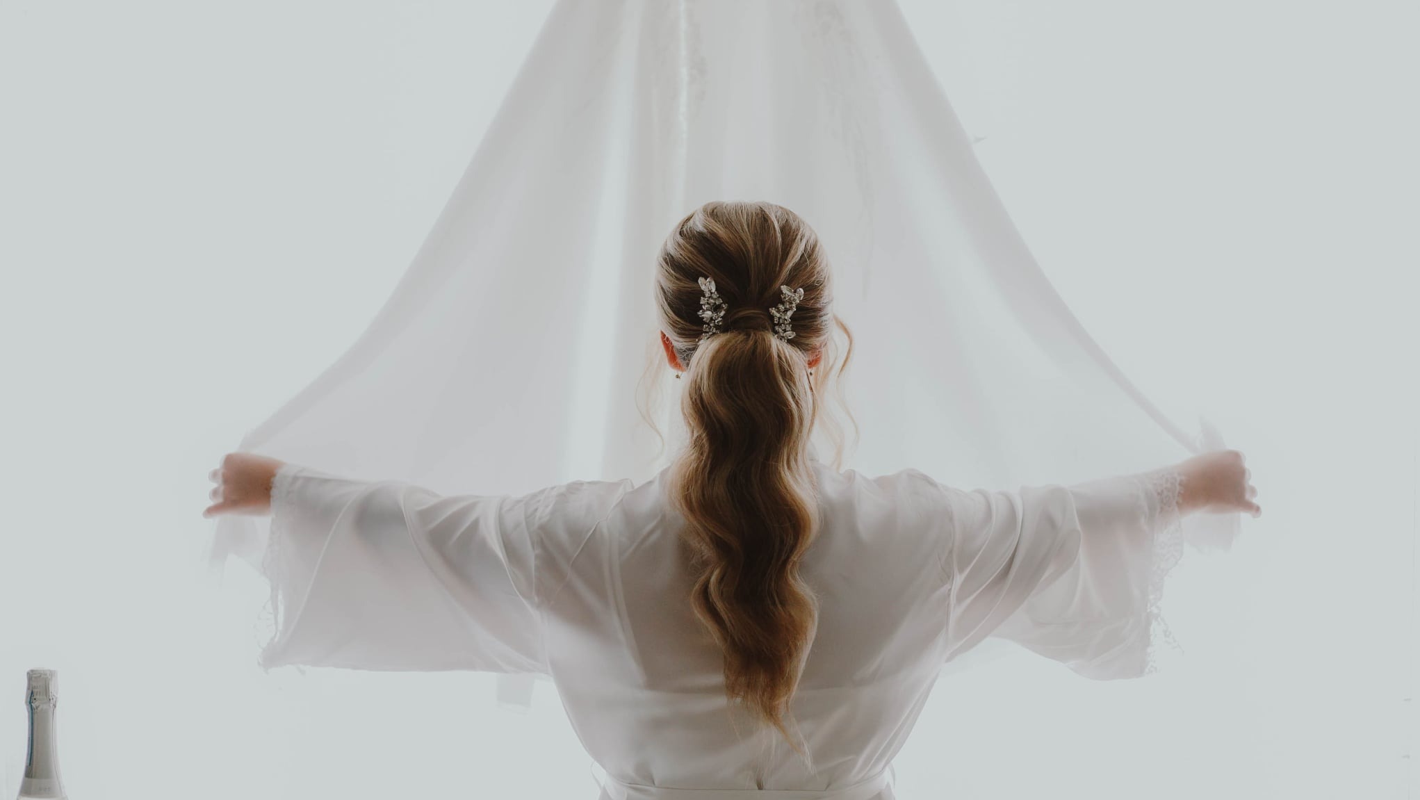 Panna młoda w szlafroku trzymająca welon na tle okna, widok z tyłu, upięte włosy z ozdobami, butelka szampana w lewym dolnym rogu.