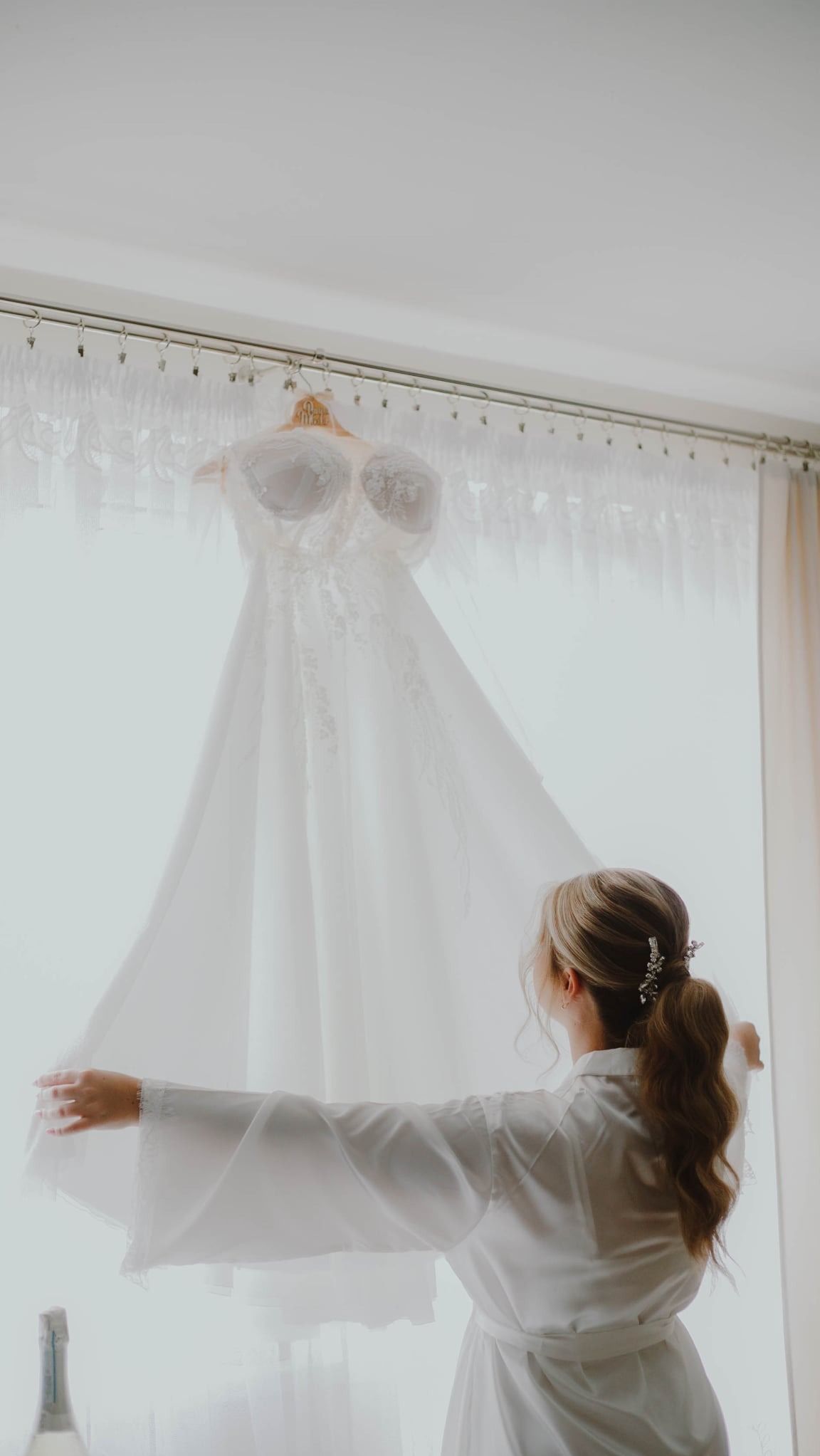 Panna młoda w białym szlafroku podziwia wiszącą suknię ślubną z koronkowym gorsetem, oświetloną przez okno z delikatnymi firankami.