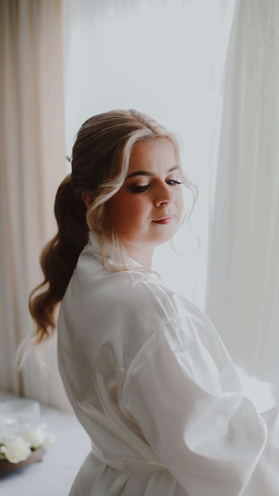 Panna młoda w białym szlafroku, z upiętymi blond włosami, spogląda w dół przy oknie. Delikatny makijaż, romantyczna fryzura, przygotowania do ślubu.