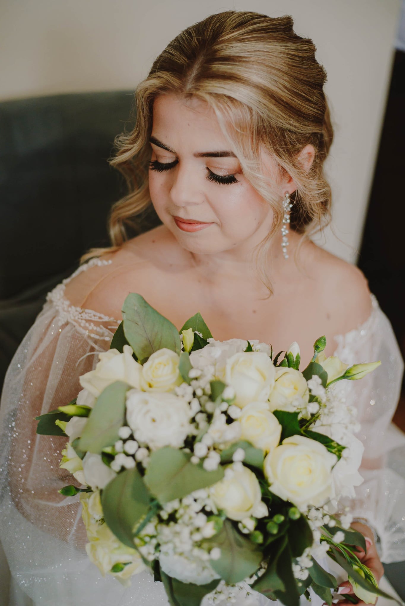 Panna młoda w sukni ślubnej z bukietem białych róż i gipsówki, spogląda w dół, widoczne kolczyki i delikatny makijaż.