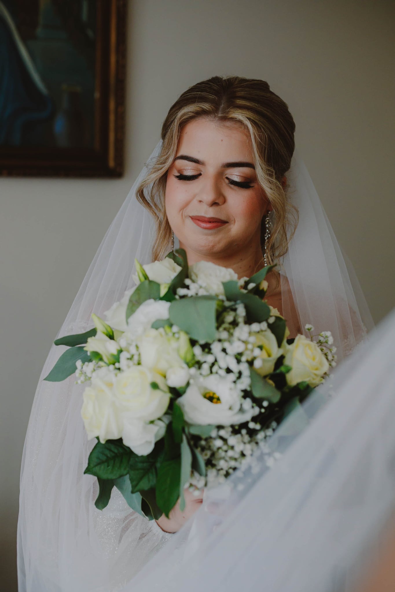 Panna młoda z bukietem białych róż i gipsówki, w welonie, z delikatnym makijażem i błyszczącymi kolczykami, spogląda w dół. W tle widoczny fragment obrazu w złotej ramie.