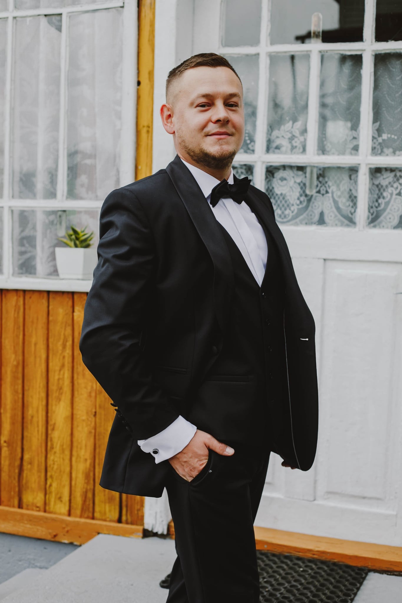 Elegancki portret mężczyzny w czarnym smokingu i muszce, pozującego na tle białych drzwi z okiennicami i drewnianego panelu.