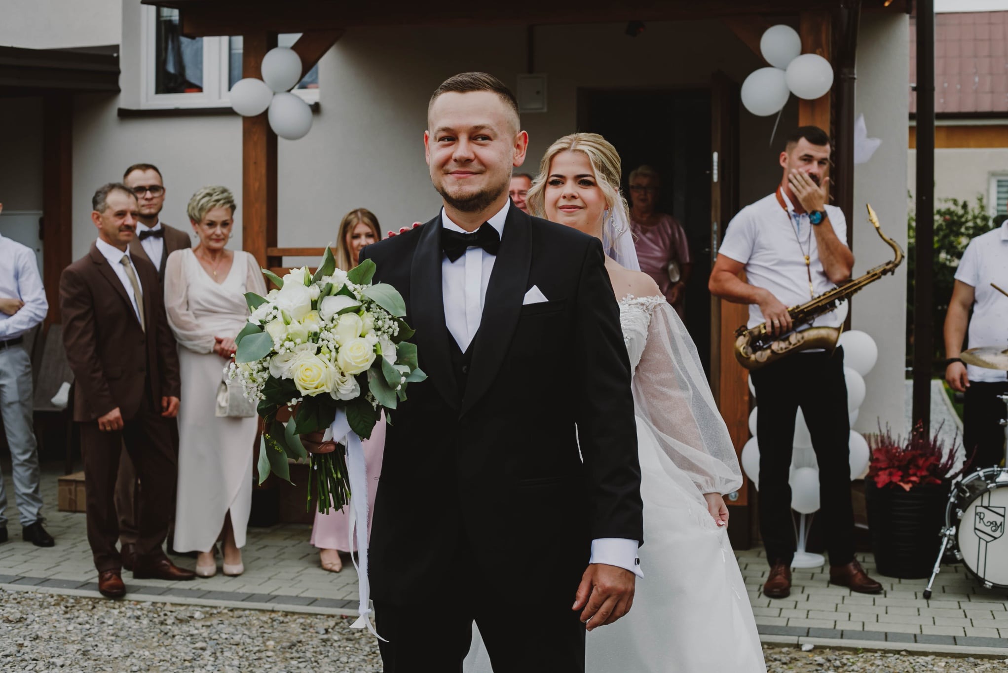 Uśmiechnięty pan młody w smokingu z bukietem, obok panna młoda w białej sukni. W tle goście, muzycy z saksofonem i perkusją. Dekoracja z białych balonów.