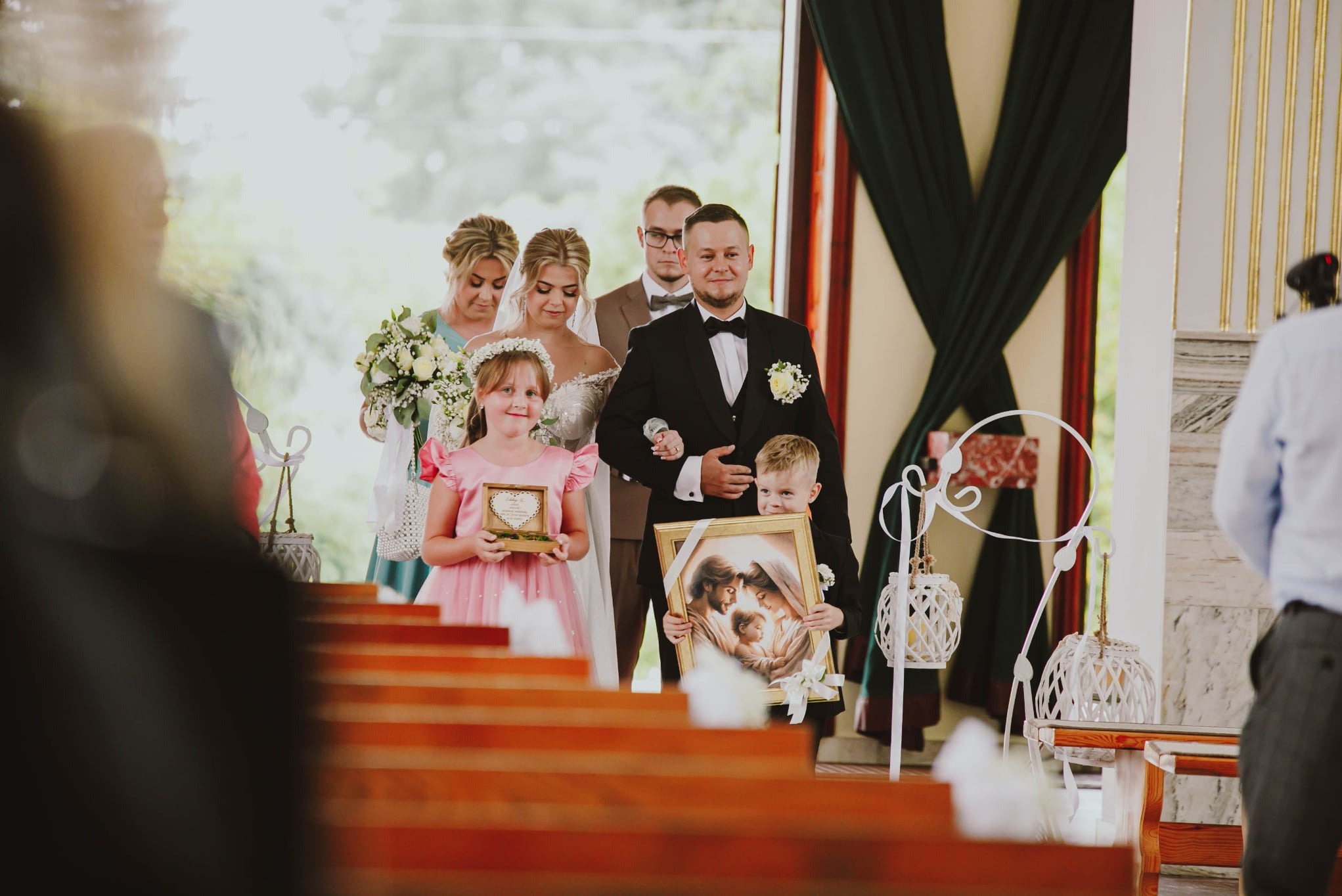 Uśmiechnięta para młoda w kościele, prowadzona przez dzieci niosące relikwie i obraz z wizerunkiem świętej rodziny. Za nimi drużba i druhna.