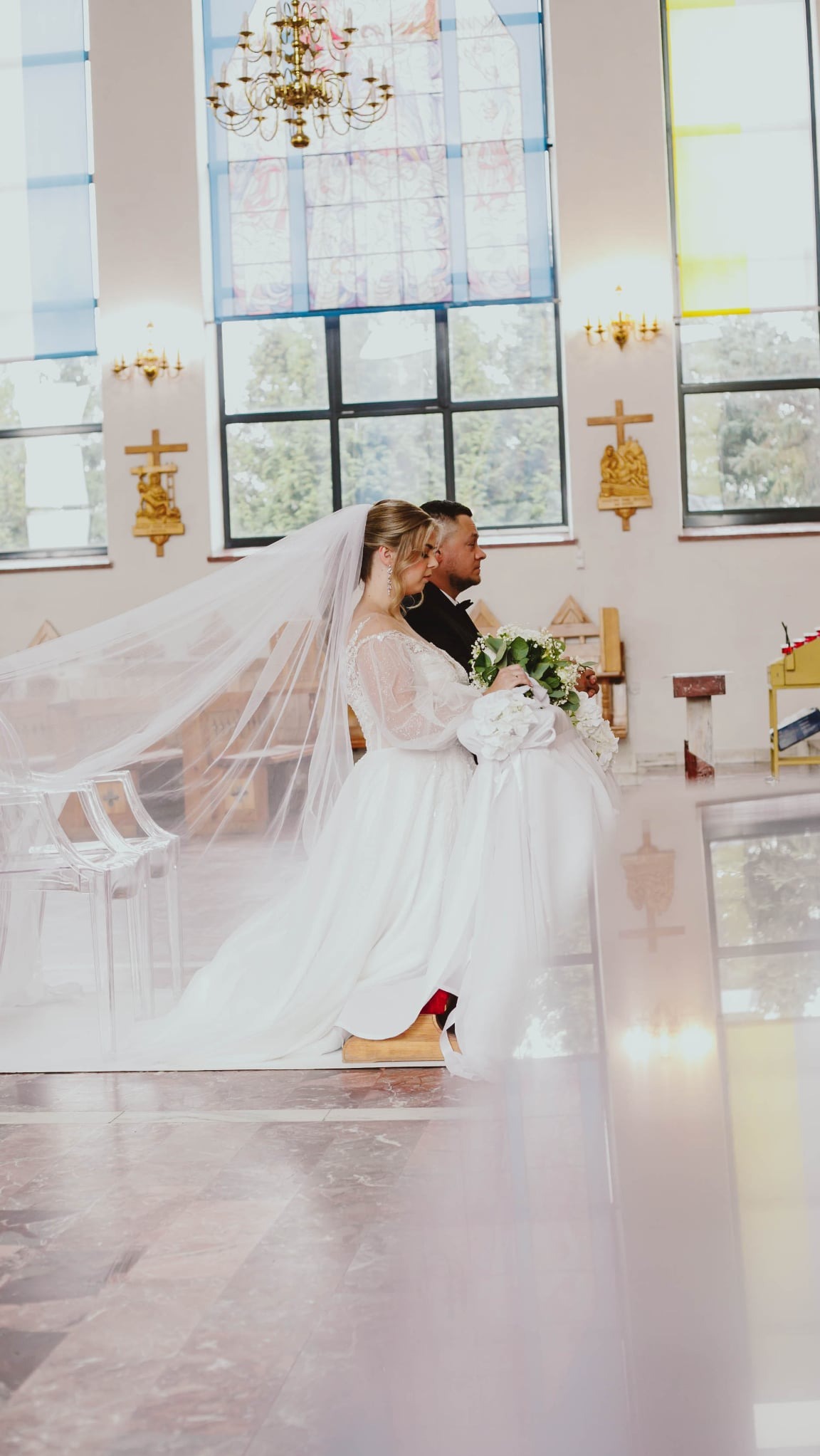 Panna młoda w białej sukni z welonem idzie do ołtarza w kościele, trzymając bukiet kwiatów. Za nią stoi mężczyzna w czarnym garniturze. W tle okna i dekoracje.