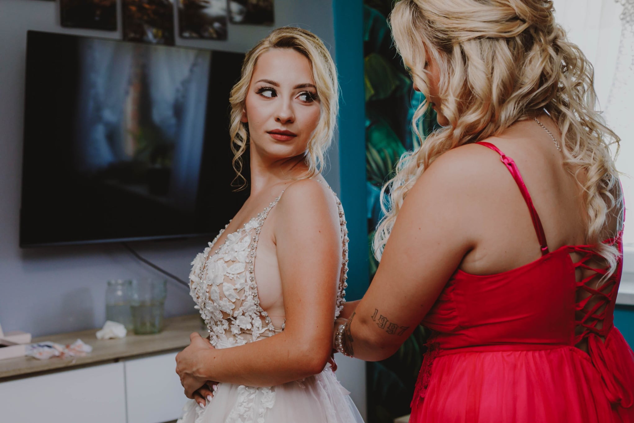 Panna młoda w koronkowej sukni ślubnej, poprawiana przez druhnę w czerwonej sukni, w pokoju z telewizorem i dekoracjami. Przygotowania do ślubu w trakcie sesji.