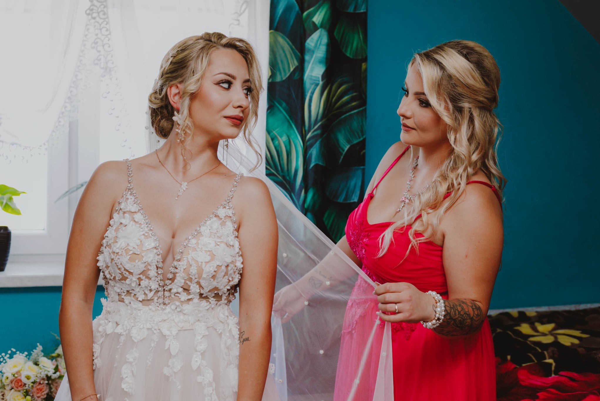 Panna młoda w koronkowej sukni spogląda na kobietę w różowej sukni, która układa jej welon. W tle widoczne są kwiaty i wzorzysta ściana.