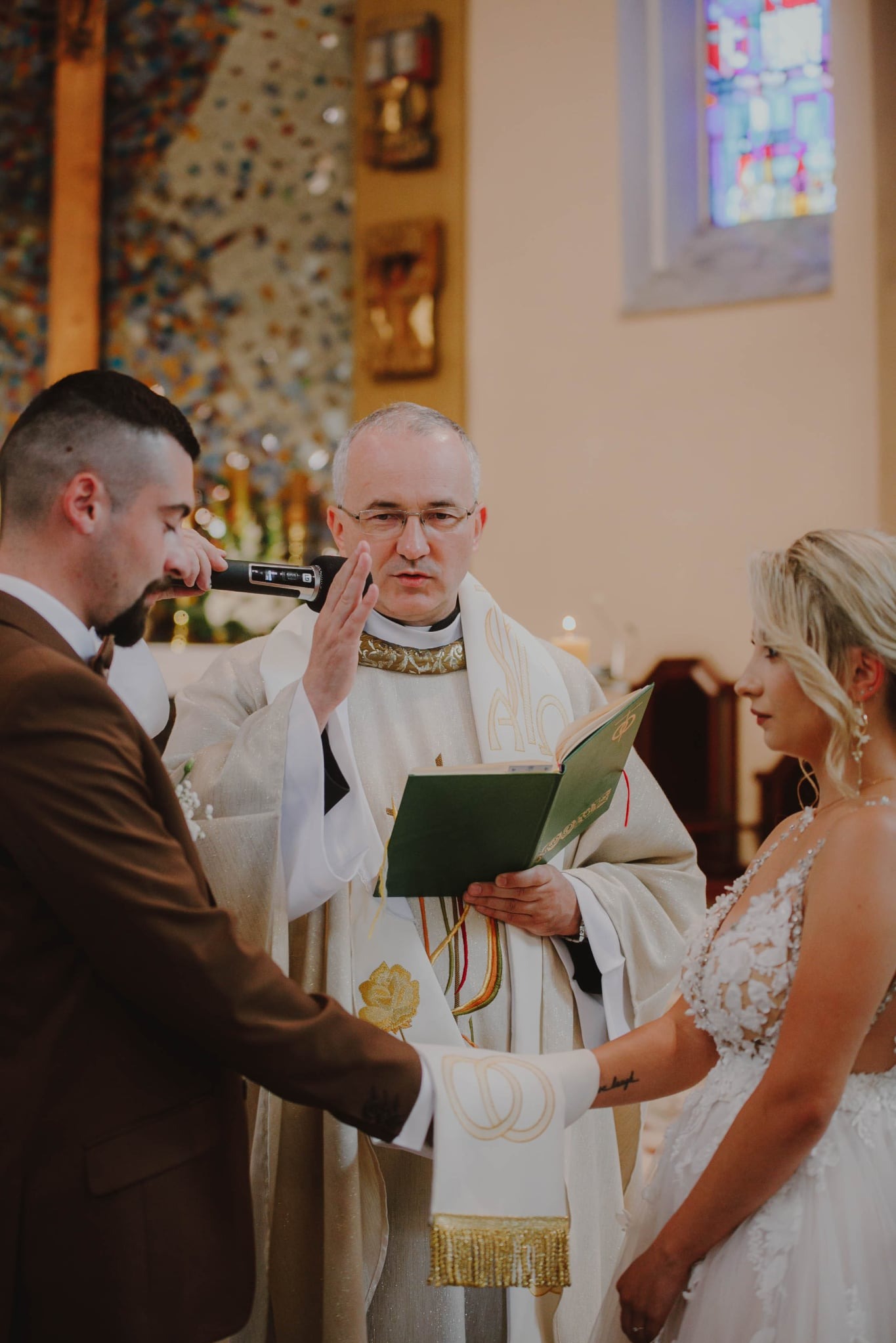 Uroczystość ślubna w kościele: Para młoda trzyma się za ręce, ksiądz błogosławi z mikrofonem. W tle witraże i dekoracje kościelne. Delikatne, naturalne światło.