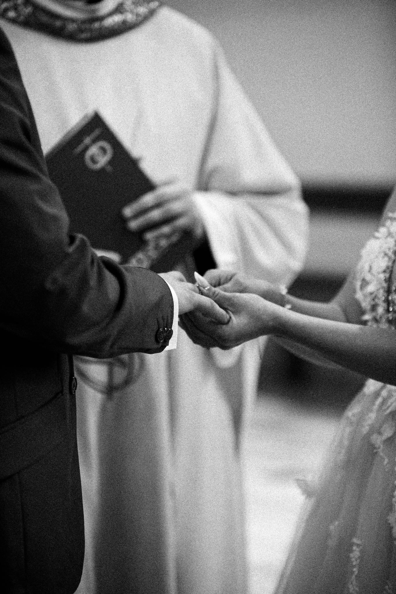 Czarno-białe zbliżenie dłoni nowożeńców podczas ceremonii ślubnej, wymiana obrączek, w tle ksiądz z księgą.