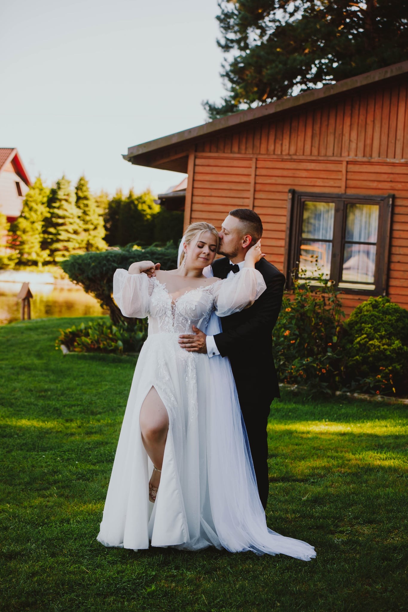 Elegancka para młoda w objęciach na tle drewnianego domu i zieleni. Panna młoda w sukni z rozcięciem, pan młody w czarnym smokingu.