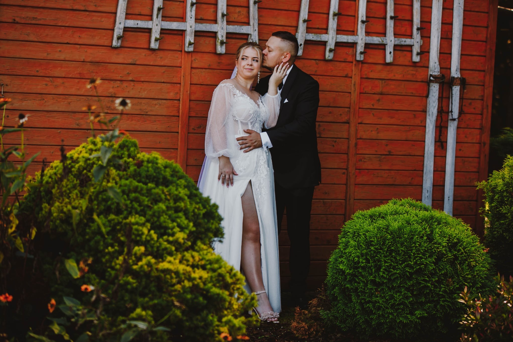 Elegancka sesja ślubna: Panna młoda w sukni z rozcięciem, oblubieniec w czarnym garniturze, czuły pocałunek na tle rustykalnej, drewnianej ściany i zieleni krzewów.
