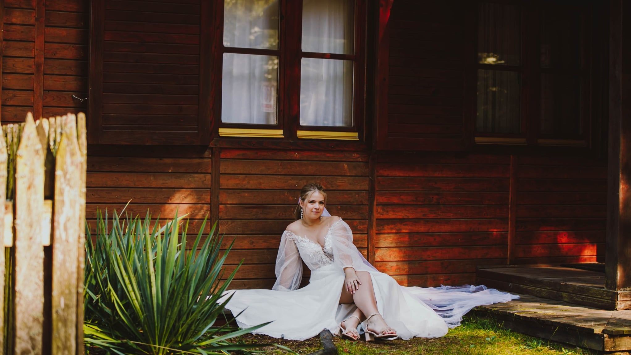 Panna młoda w zwiewnej sukni siedzi na ziemi przed drewnianym domkiem, z widoczną zielenią i fragmentem płotu. Ujęcie z bliska, ciepłe światło.