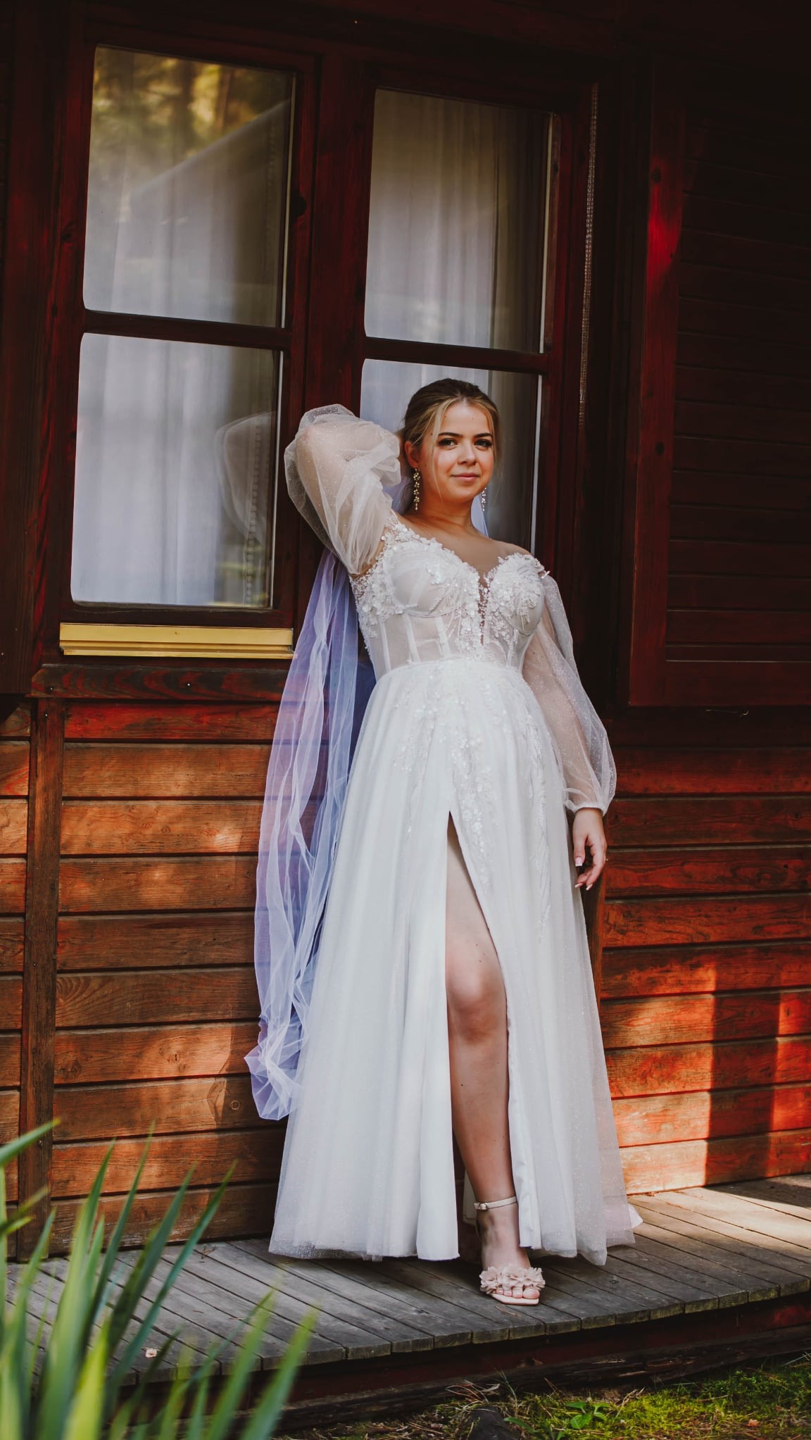 Panna młoda w koronkowej sukni z rozcięciem, pozuje na tle drewnianego domu z oknem. Delikatny welon i ozdobne kolczyki dopełniają stylizację.