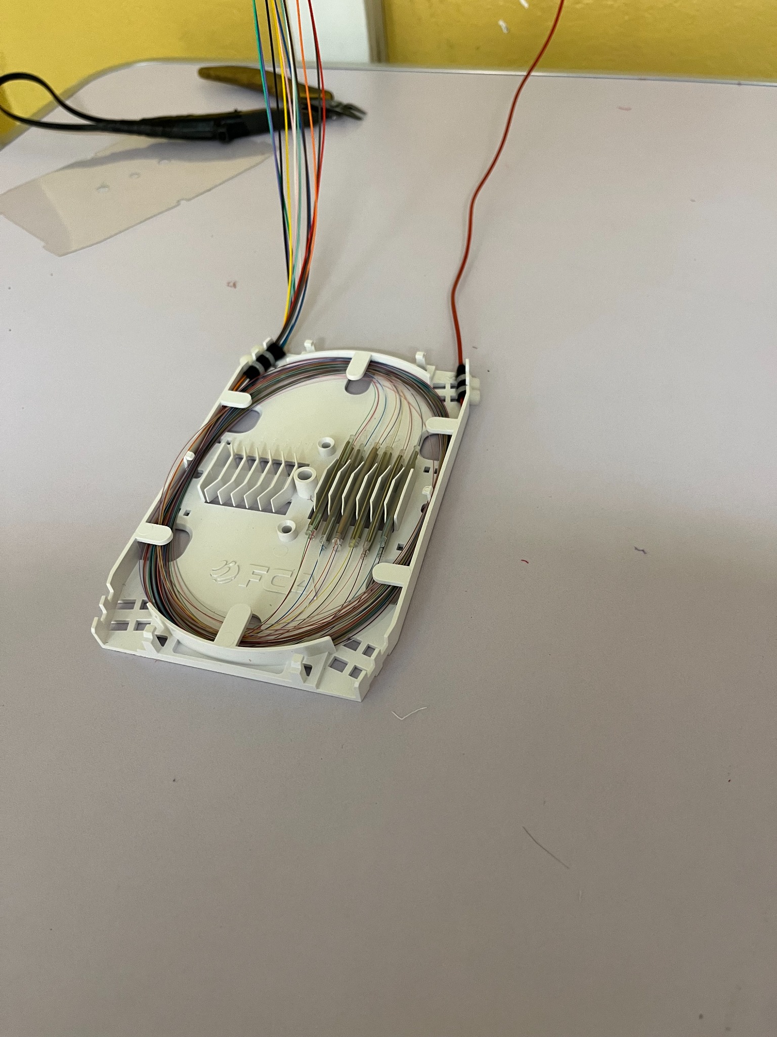 Rozdzielnica światłowodowa z widocznymi włóknami w różnych kolorach, ułożonymi wewnątrz białej obudowy. Obok leżą szczypce do cięcia kabli.