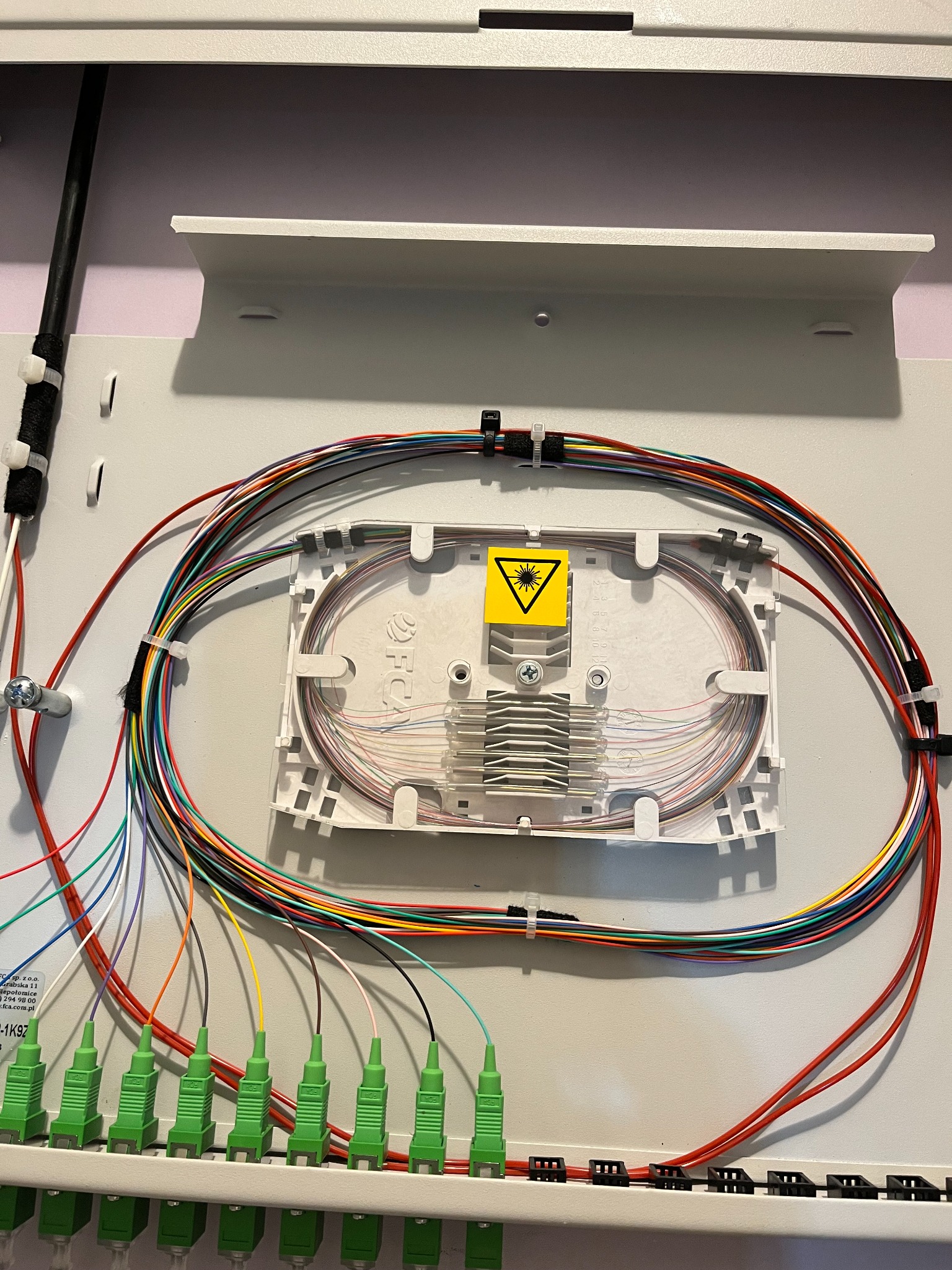 Zamknięta skrzynka światłowodowa z kolorowymi kablami i żółtą naklejką ostrzegawczą. Precyzyjne ułożenie włókien wewnątrz. Estetyczne wykończenie instalacji.