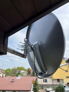 Zamontowana antena satelitarna na balkonie z widokiem na zabudowę jednorodzinną i pochmurne niebo. Antena przymocowana do metalowej konstrukcji.