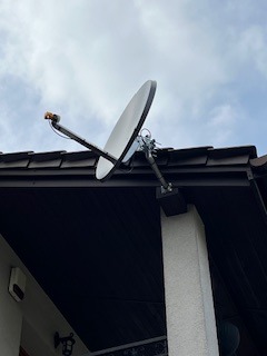 Antena satelitarna zamontowana na dachu budynku, widoczna z dołu pod kątem, na tle pochmurnego nieba. Brązowa dachówka i biała kolumna wspierają konstrukcję.