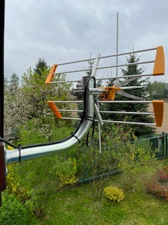 Antena Yagi-Uda z pomarańczowymi elementami, zamontowana na balkonie, widok z boku na tle zieleni. Kabel antenowy poprowadzony wzdłuż metalowego masztu.