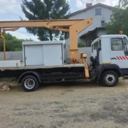 Biała ciężar&oacute;wka z ż&oacute;łtym dźwigiem na podw&oacute;rku, obok śpi pies. W tle dom i drzewa. Widok z boku, w dzień.