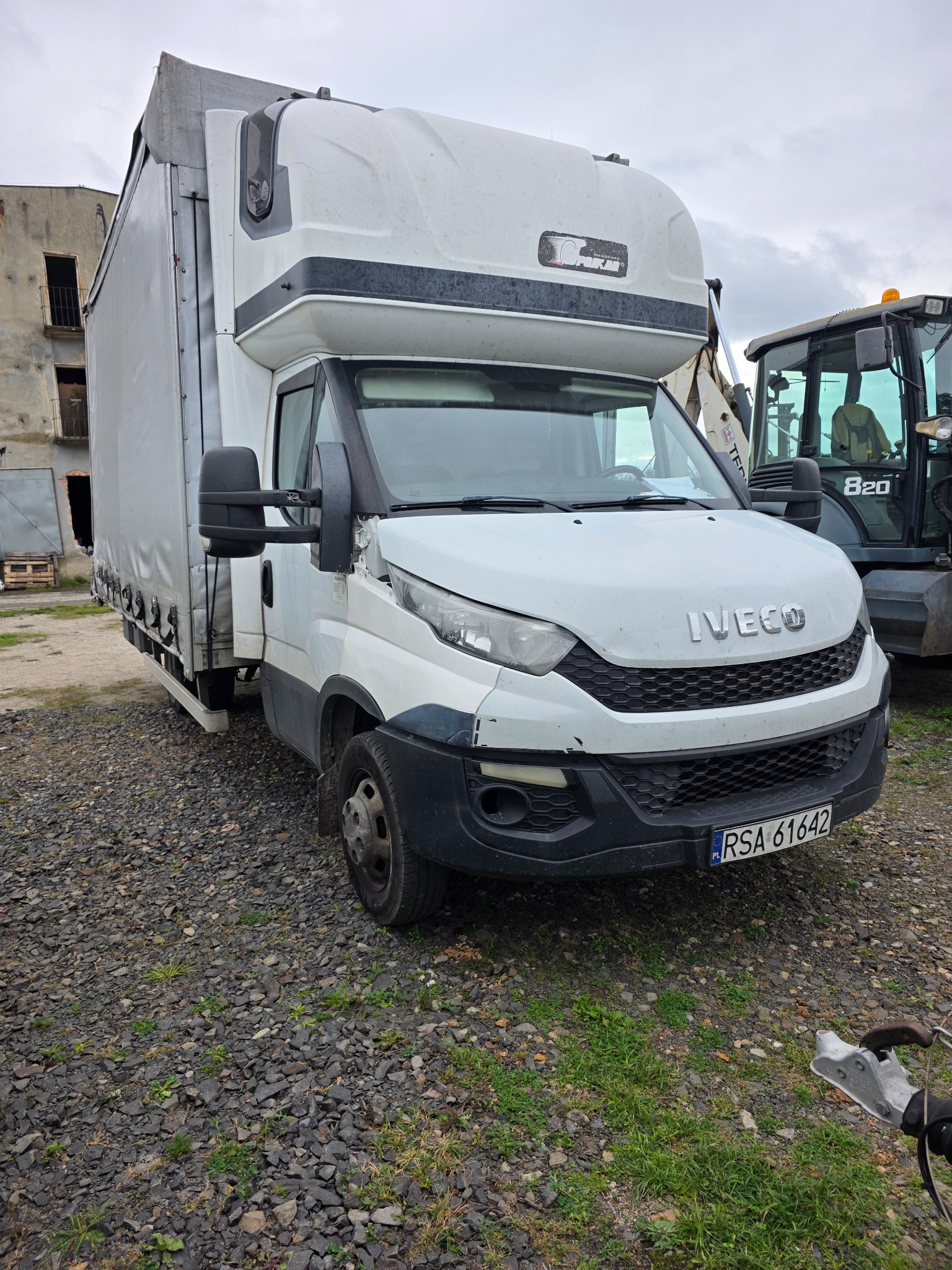 Biały samochód dostawczy Iveco z plandeką, zaparkowany na żwirowym podłożu. W tle budynek i maszyna budowlana. Widoczne tablice rejestracyjne.