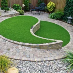 Ogród z trawnikiem w kształcie yin yang, obrzeża z kostki brukowej i kamieni, ozdobne rośliny, meble ogrodowe w tle, dekoracyjna figurka.