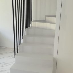KS MIKROCEMENT Karol Specjał - Białe schody z mikrocementu z czarną balustradą na tle białych ścian i marmurowej podłogi. Minimalistyczny design.