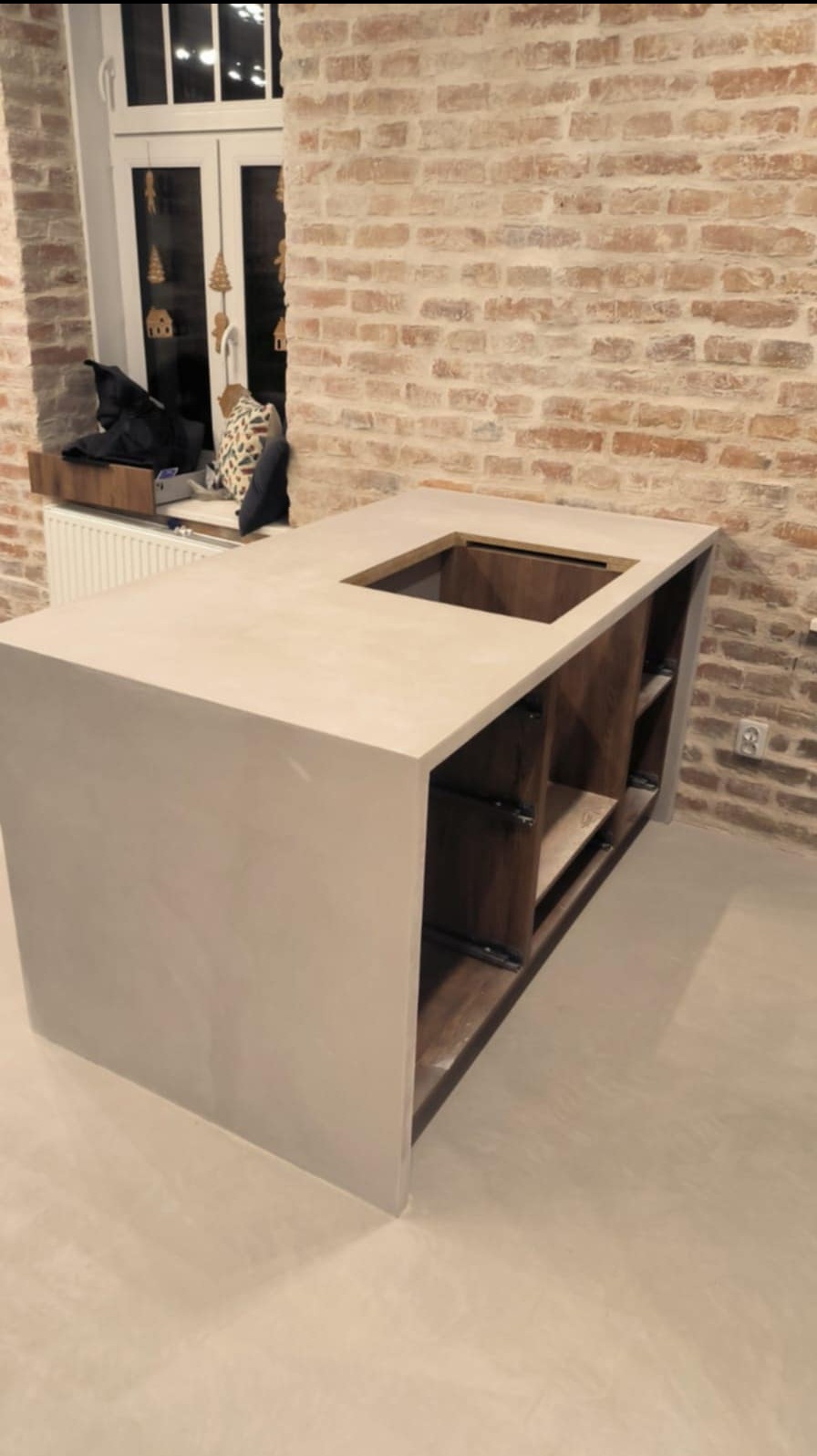 Wykończona wyspa kuchenna z blatem z materiału imitującego beton, z otworem na zlew, na tle ściany z czerwonej cegły i okna.