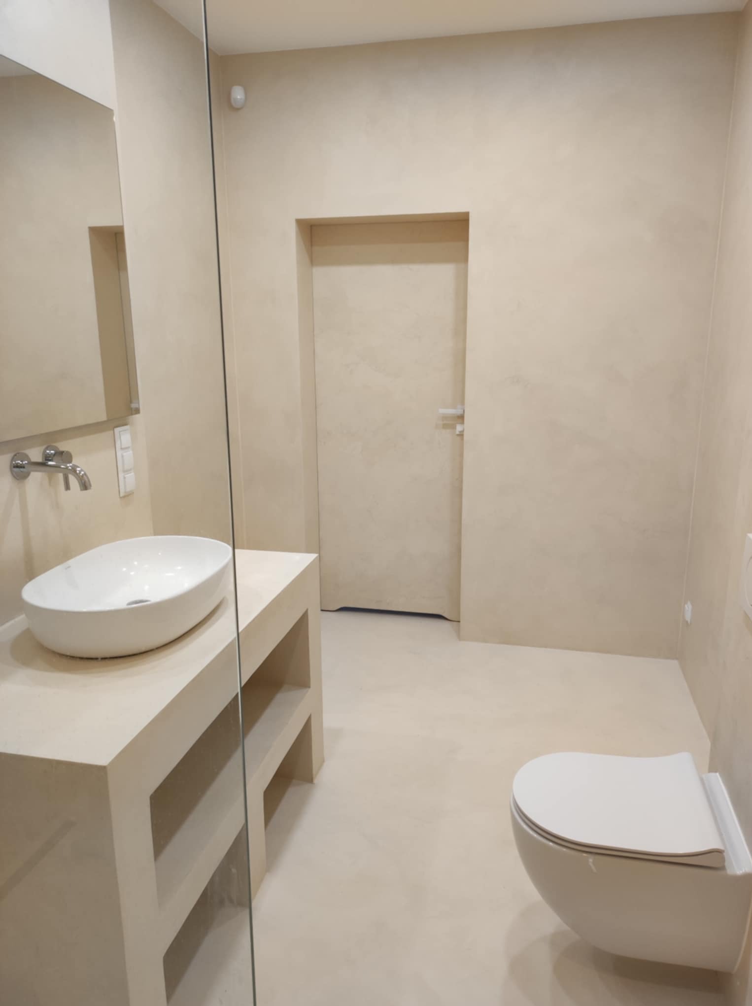 Beżowa łazienka z umywalką nablatową i wiszącą toaletą, wykończona jednolitą powierzchnią ścian i podłogi, z drzwiami w tle.