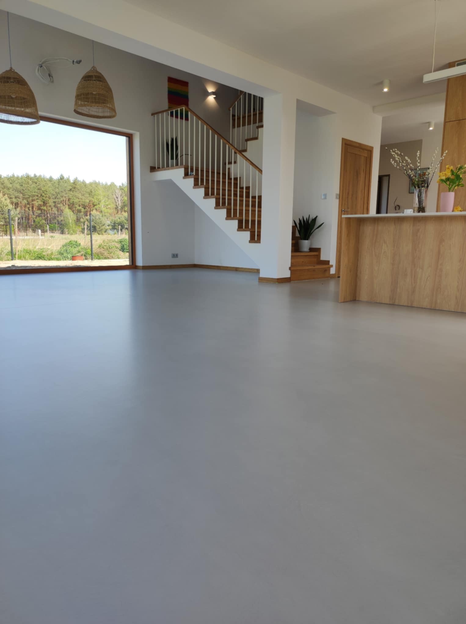 Gładka, szara posadzka w jasnym wnętrzu domu w Trzcince. Widoczne schody, duże okno z widokiem na zieleń i fragment drewnianej zabudowy kuchennej.