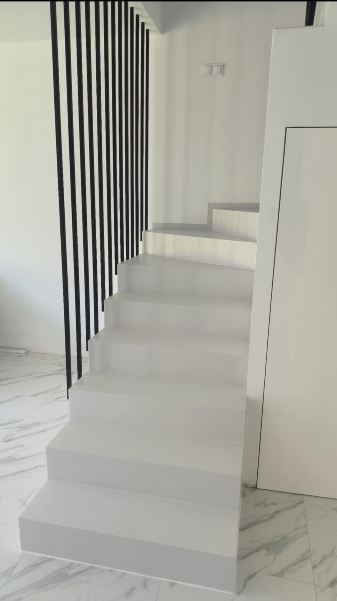Białe schody z mikrocementu z czarną balustradą na tle białych ścian i marmurowej podłogi. Minimalistyczny design.