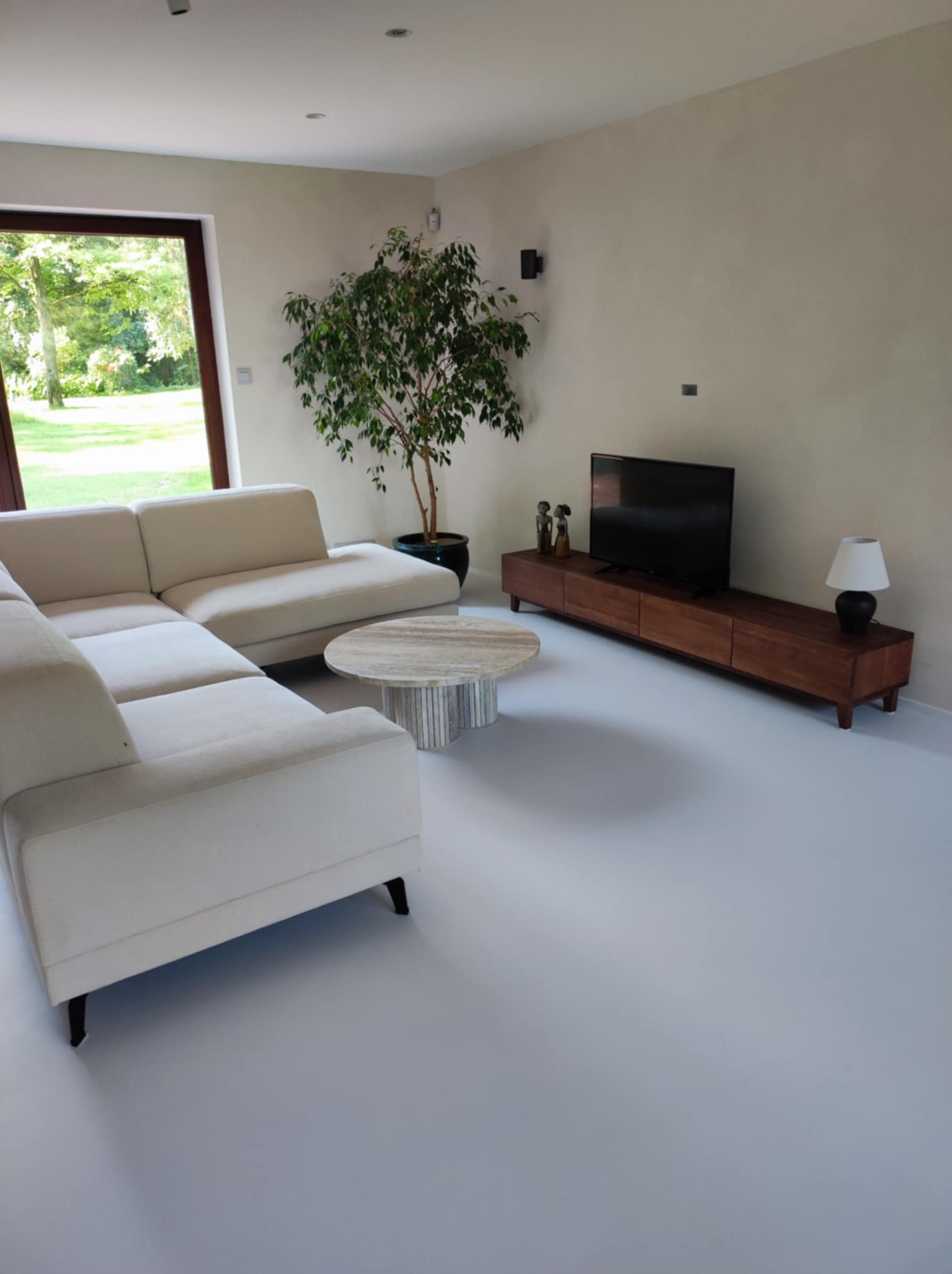 Jasnoszara, gładka posadzka w salonie z narożnikiem, stolikiem kawowym i szafką RTV. Duże okno z widokiem na zieleń. Minimalistyczny wystrój.