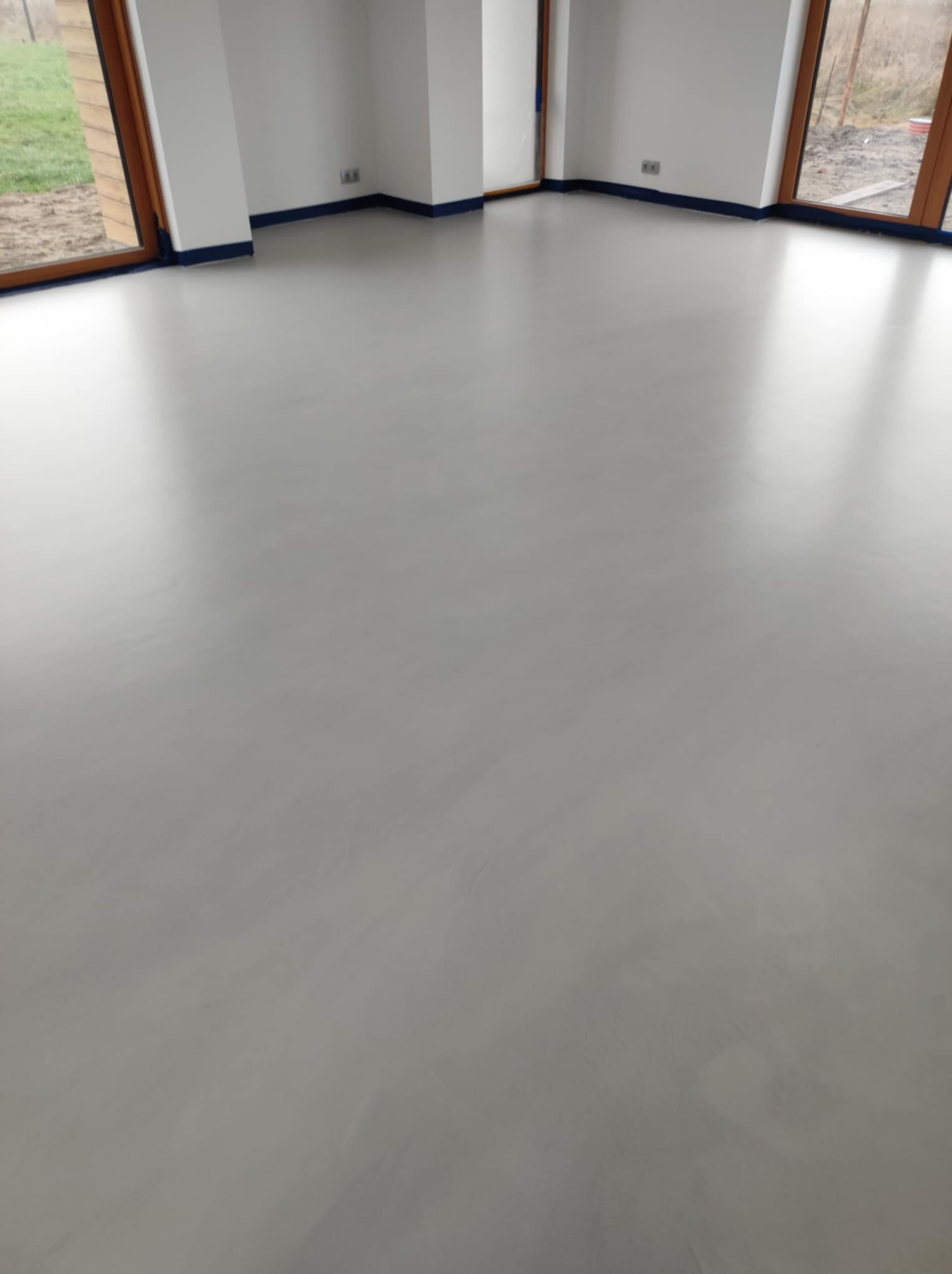 Jasnoszara, gładka posadzka w nowym pomieszczeniu z oknami i zabezpieczonymi listwami przypodłogowymi. Minimalistyczny design, świeżo po realizacji.