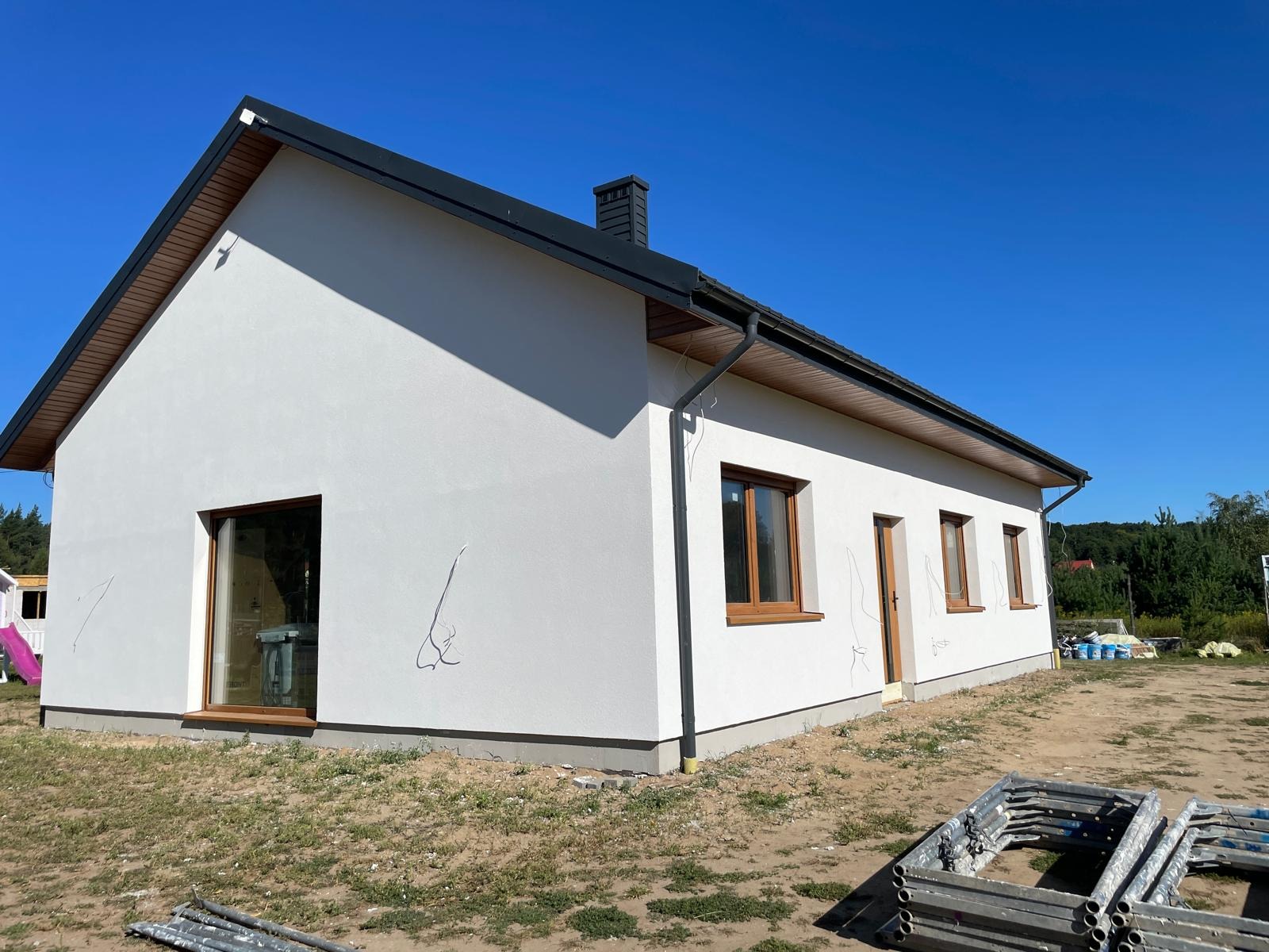 Biały dom jednorodzinny z grafitowym dachem i drewnianymi oknami. Na ścianie widoczne ślady po dociepleniu. Na ziemi leżą rusztowania. Błękitne niebo w tle.