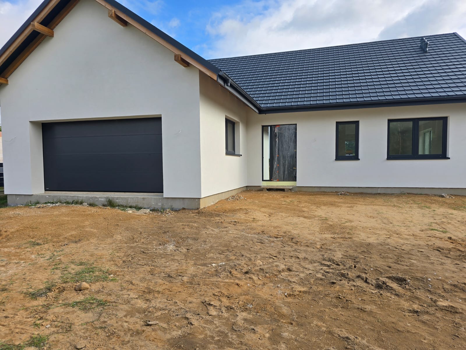 Biały dom jednorodzinny z czarną bramą garażową i stolarką okienną. Elewacja wykończona, teren wokół domu przygotowany pod zagospodarowanie.