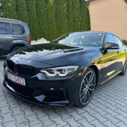 Lśniący, czarny samochód BMW serii 4 Gran Coupe z widoczną tablicą rejestracyjną, zaparkowany na brukowanym podjeździe przed żywopłotem.