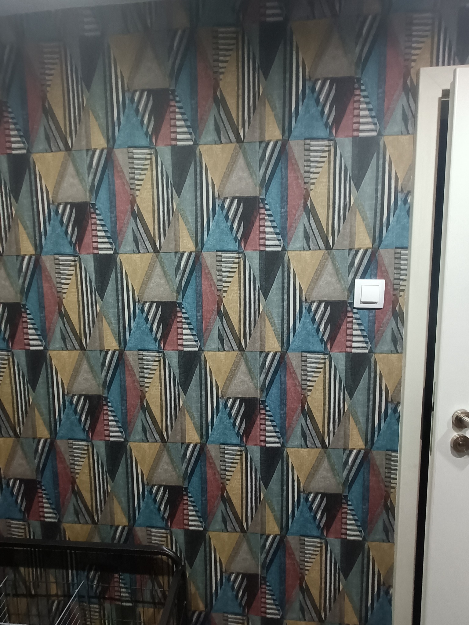 Wzorzysta tapeta w geometryczne trójkąty w odcieniach niebieskiego, żółtego, czerwonego i szarości. Widoczny biały włącznik światła i fragment białych drzwi.
