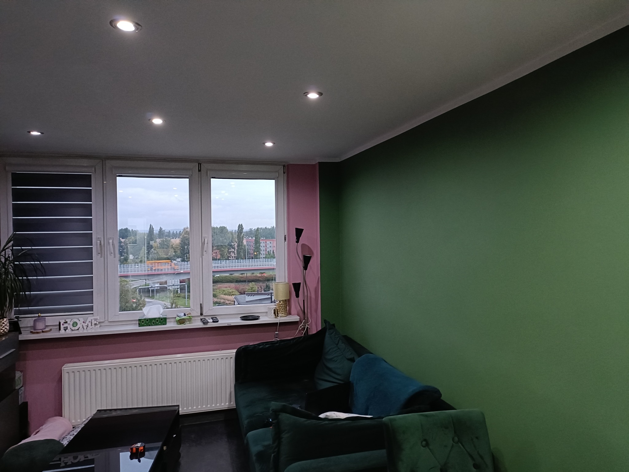Salon po malowaniu ścian na zielono i różowo, z ciemnozieloną sofą i widokiem z okna na miasto. Nowoczesne oświetlenie sufitowe.