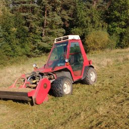 Przedsiębiorstwo Transportowo - Usługowo - Handlowe RES-STAN Stanisław Reszkiewicz - Czerwony traktor z kosiarką listwową na nierównym terenie z niską trawą. W tle drzewa i krzewy w słoneczny dzień.