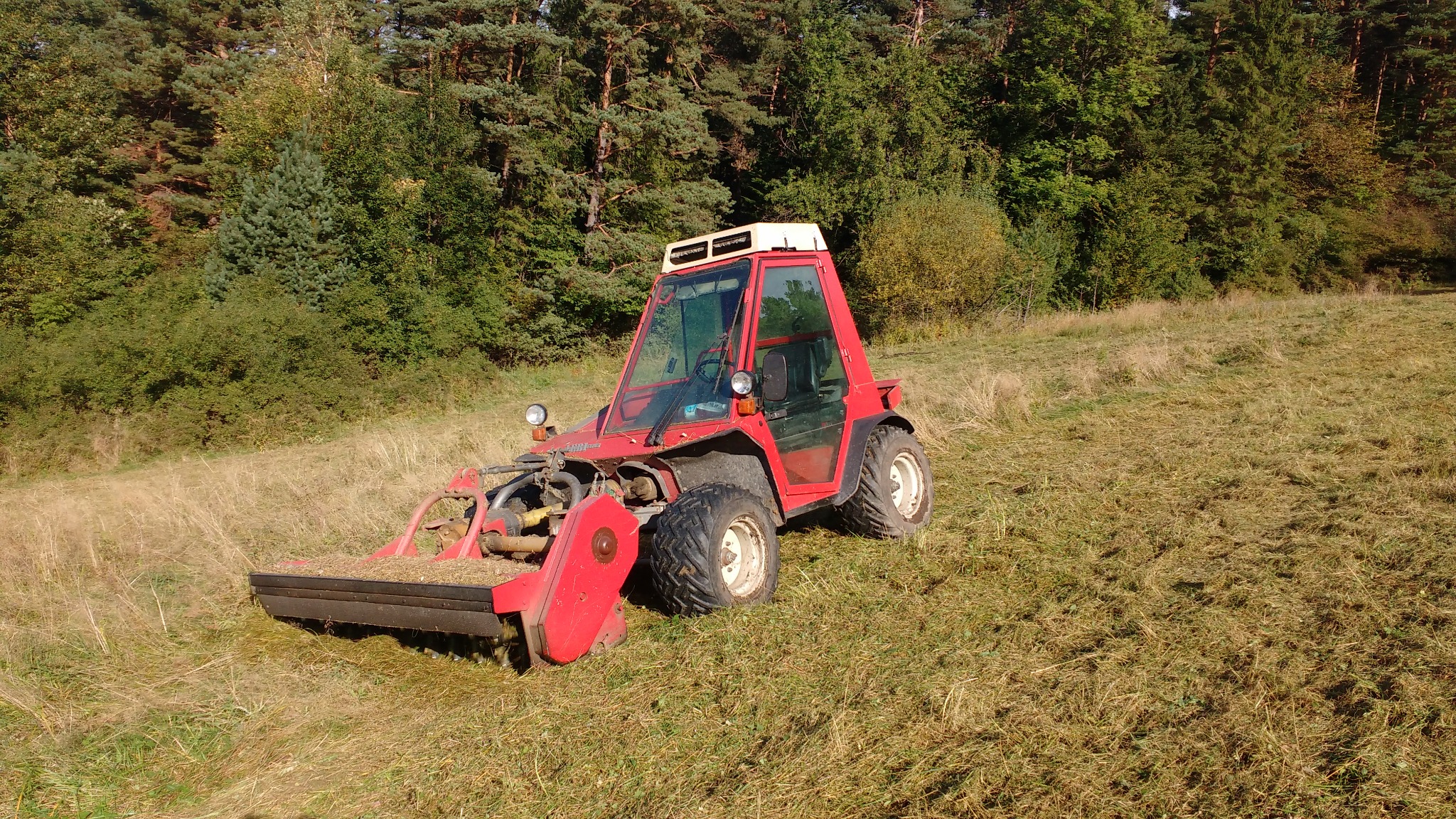 Czerwony traktor z kosiarką listwową na nierównym terenie z niską trawą. W tle drzewa i krzewy w słoneczny dzień.