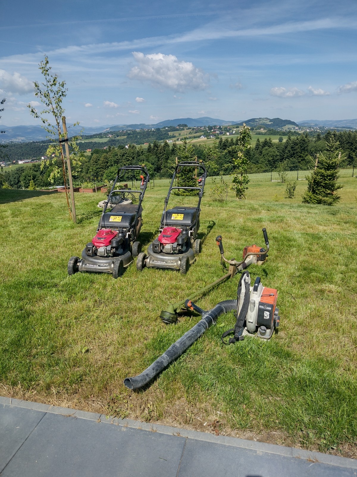 Dwie kosiarki Honda i dmuchawa do liści na trawniku z widokiem na pagórkowaty krajobraz w tle, w słoneczny dzień. Sprzęt ogrodniczy.