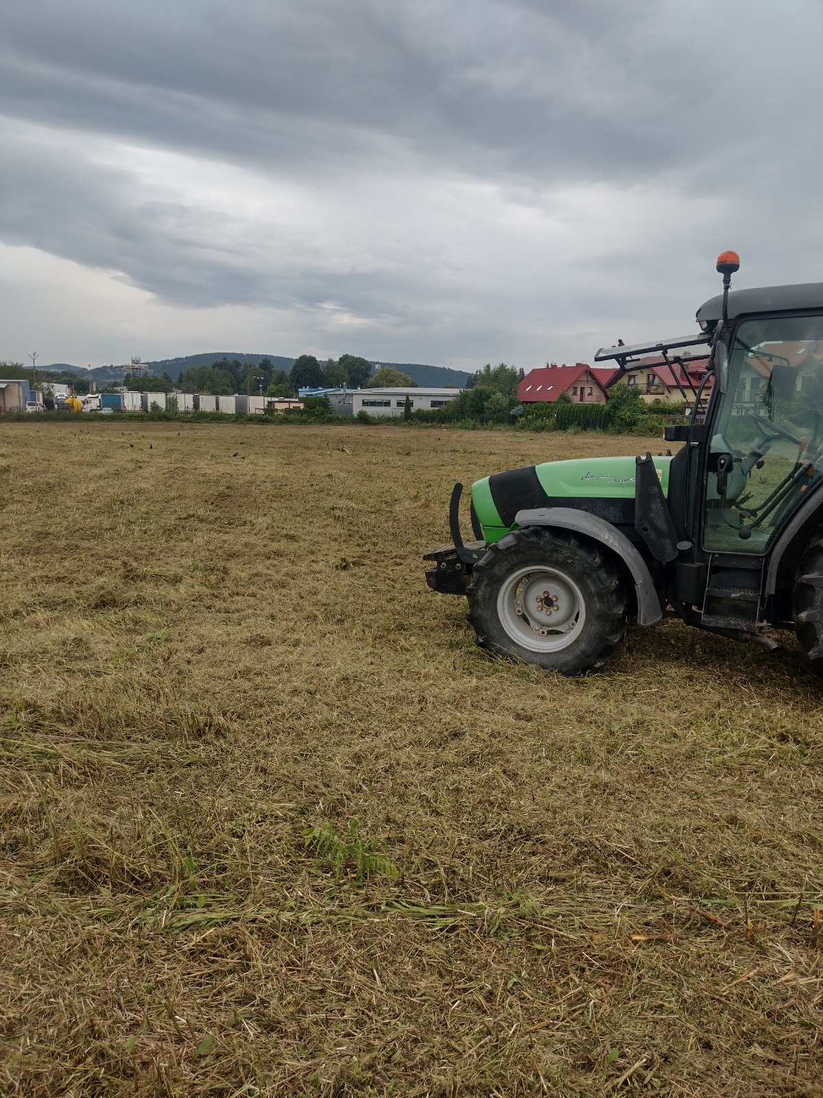 Zielony traktor Deutz-Fahr na polu po koszeniu trawy. Ściernisko, w tle budynki i wzgórza pod pochmurnym niebem. Widok z boku na maszynę.