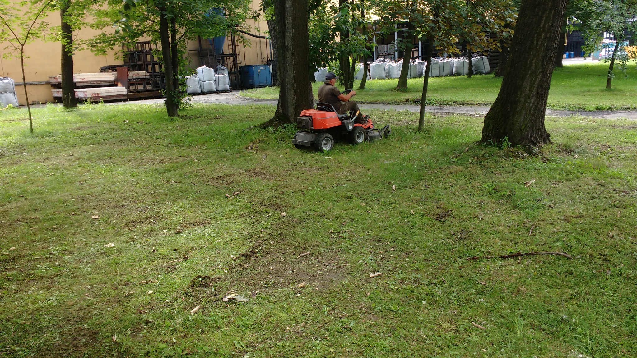 Mężczyzna kosi trawę traktorkiem Husqvarna na zielonym trawniku z drzewami. Ślady po koszeniu widoczne na trawie.