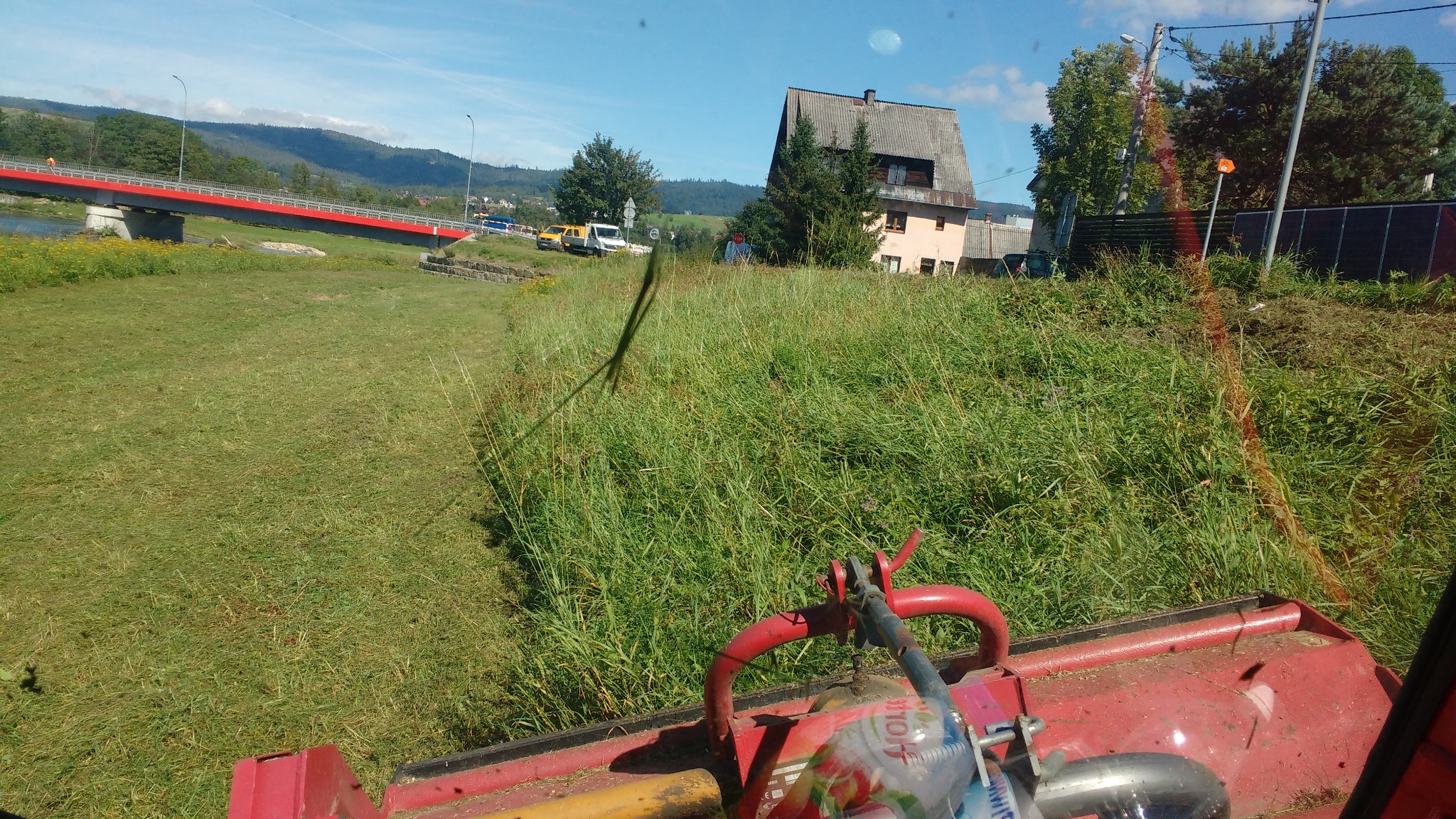 Traktor koszący trawę na łące z widokiem na czerwony most i budynki w tle. Ujęcie z perspektywy operatora maszyny.