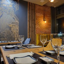 MEGA CODE ADAM CZEMKO - Stylowa aranżacja wnętrza restauracji z motywami azjatyckimi: dekoracyjna ściana z rysunkami, ceglana ściana, drewniane lamele i nakryty stół z zastawą.